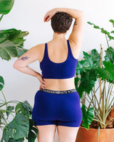 Rear view portrait of a model showcasing the scoop back fit of the deep blue Thunderpants US Indigo Montoya Longline Bra paired with matching branded waistband shorts.