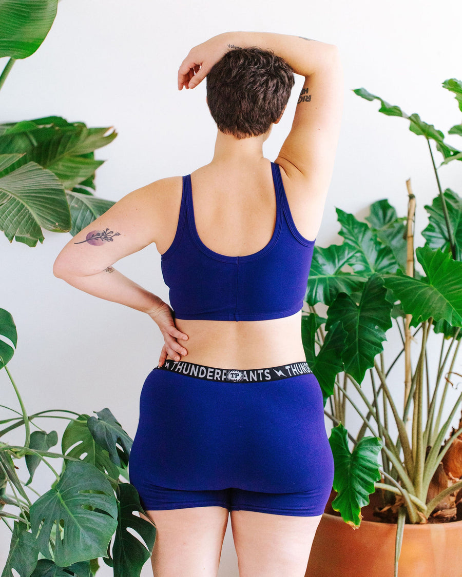 Rear view portrait of a model showcasing the scoop back fit of the deep blue Thunderpants US Indigo Montoya Longline Bra paired with matching branded waistband shorts.