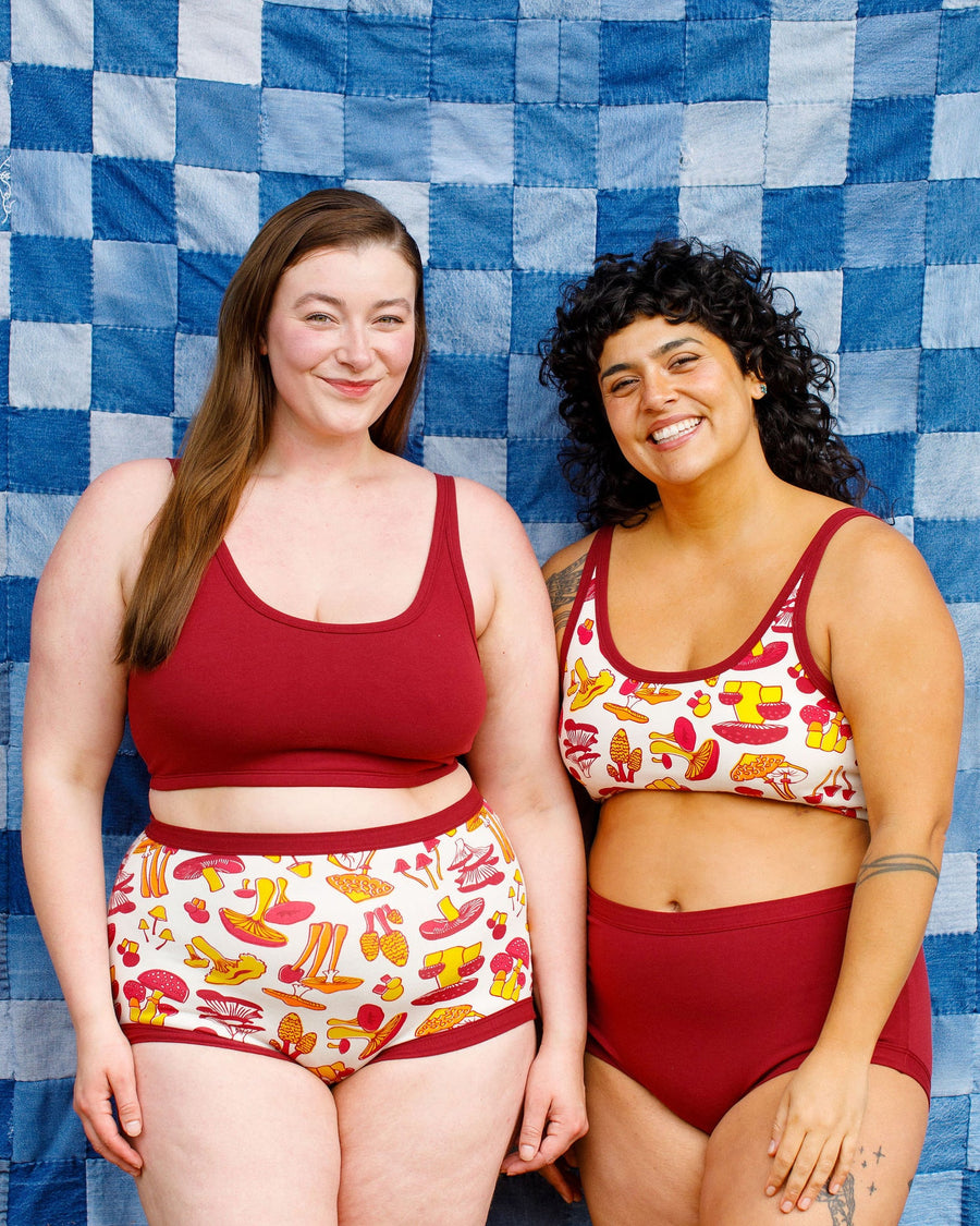 Studio portrait of two models posing together in matching Tuscan Red and mushroom print organic cotton underwear sets against a blue patchwork backdrop.