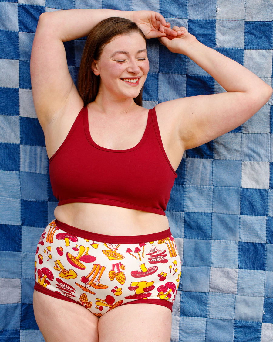 Front view of a smiling model reclining against a blue patchwork backdrop, wearing the Tuscan Red organic cotton longline bra paired with mushroom print briefs.