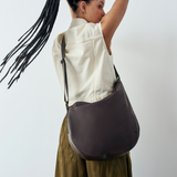 Outdoor lifestyle shot of a model with braided hair wearing the black Luna Crossbody bag with a cream vest and olive pants.