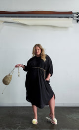 Studio lifestyle photo of a model holding a metallic woven bucket bag, wearing the Baacal black Maxine Kaftan styled with a gold chain belt.