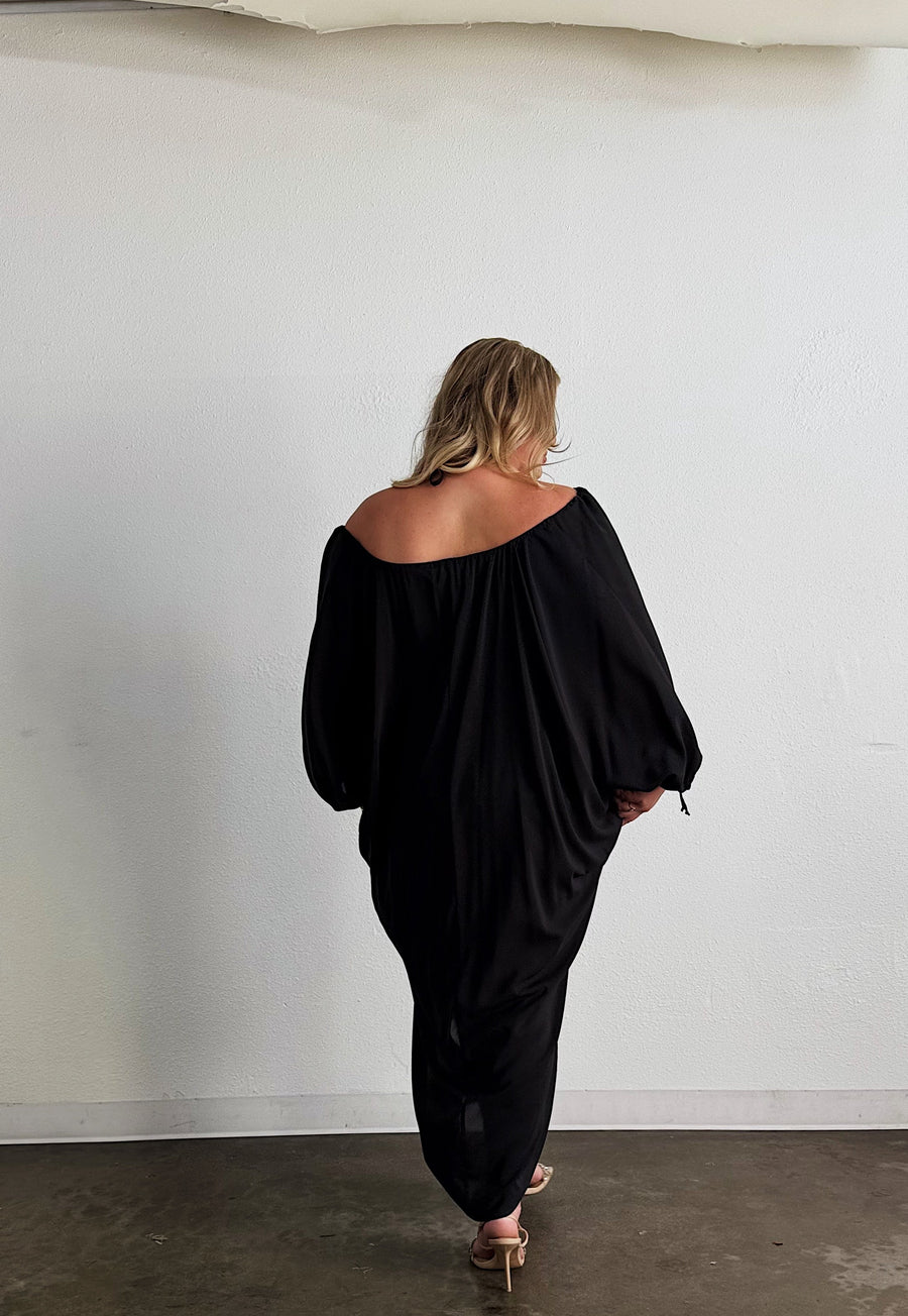 Rear view of a blonde model wearing the black Baacal Maxine Kaftan off-the-shoulder, paired with nude heel sandals.