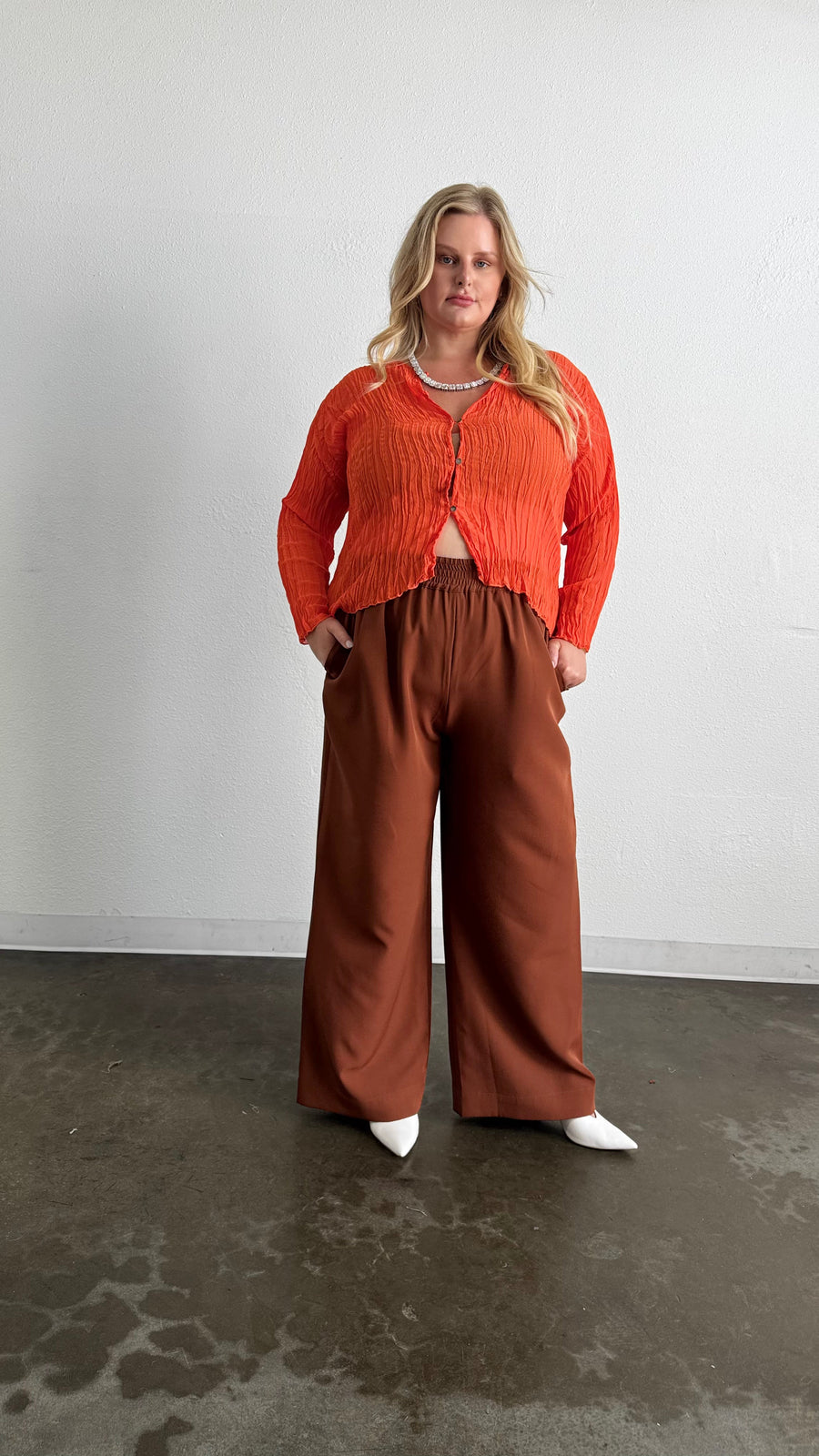 Three-quarter front portrait of a model wearing the orange Maya Pleated Cardigan buttoned at the top over brown wide-leg pants.