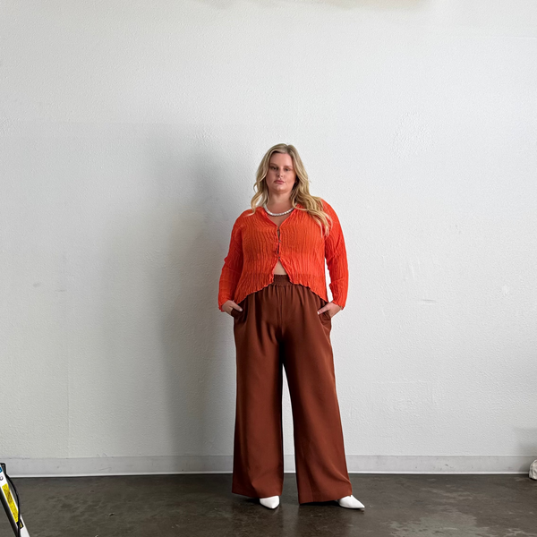 Full-length studio portrait of a blonde model wearing the orange Maya Pleated Cardigan paired with wide-leg terracotta trousers and white pointed mules.