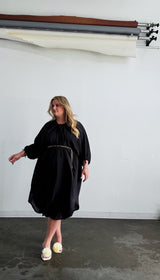 Full-length photo of a model wearing the black Maxine Kaftan belted at the waist with a thin gold chain belt and pom-pom heels.