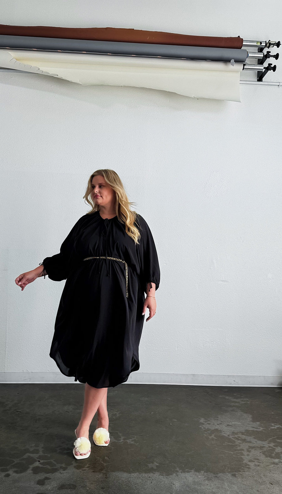 Full-length photo of a model wearing the black Maxine Kaftan belted at the waist with a thin gold chain belt and pom-pom heels.