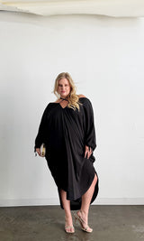 Front view of a blonde model posing in the black Baacal Maxine Kaftan styled with a gold clutch and strappy heels.