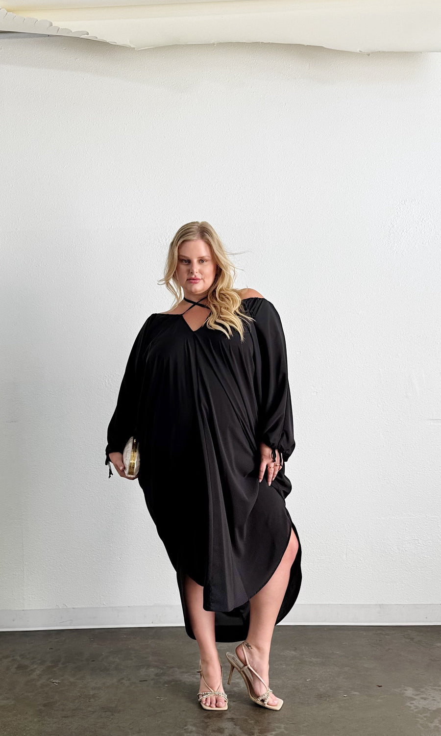 Front view of a blonde model posing in the black Baacal Maxine Kaftan styled with a gold clutch and strappy heels.