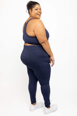 Side profile of a plus-size woman wearing a matching navy blue activewear set consisting of a sports bra and high-waisted leggings, paired with white sneakers, photographed against a solid white studio background.