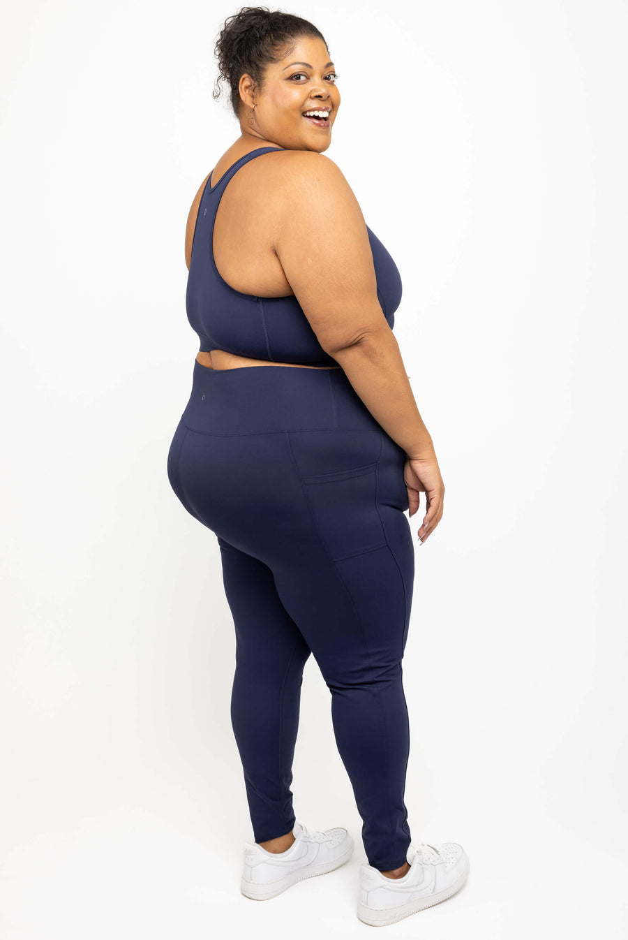 Side profile of a plus-size woman wearing a matching navy blue activewear set consisting of a sports bra and high-waisted leggings, paired with white sneakers, photographed against a solid white studio background.