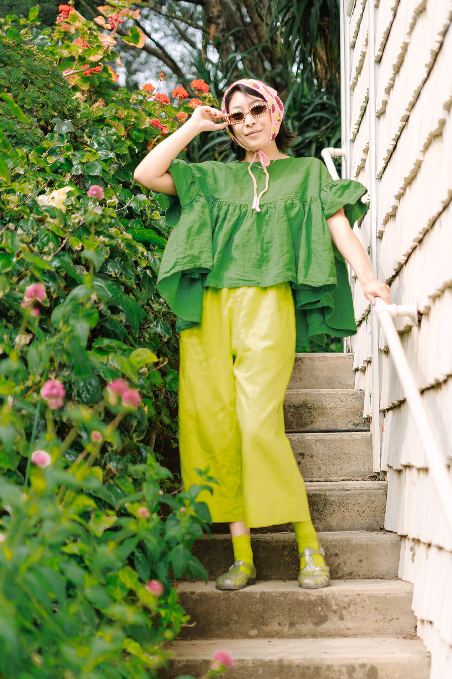 Full-length outdoor portrait of a model posing on stairs wearing Nettle Studios Limeade Linen Mid Easy Pants with a bonnet, sunglasses, and jelly shoes.