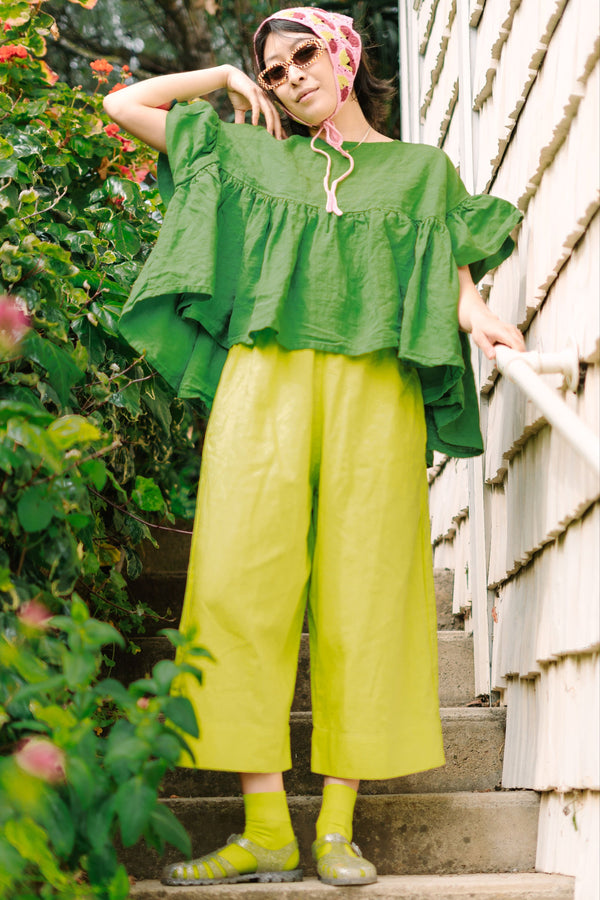 Low-angle outdoor shot of a model standing on stairs wearing size-inclusive limeade green wide-leg linen pants paired with an oversized green tiered smock top.