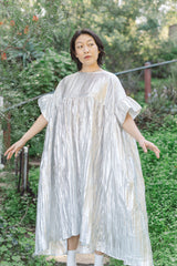 Full-length outdoor portrait of a model in a lush green garden setting wearing the silver metallic crinkle lamé mushroom dress showing its slight high-low hem.
