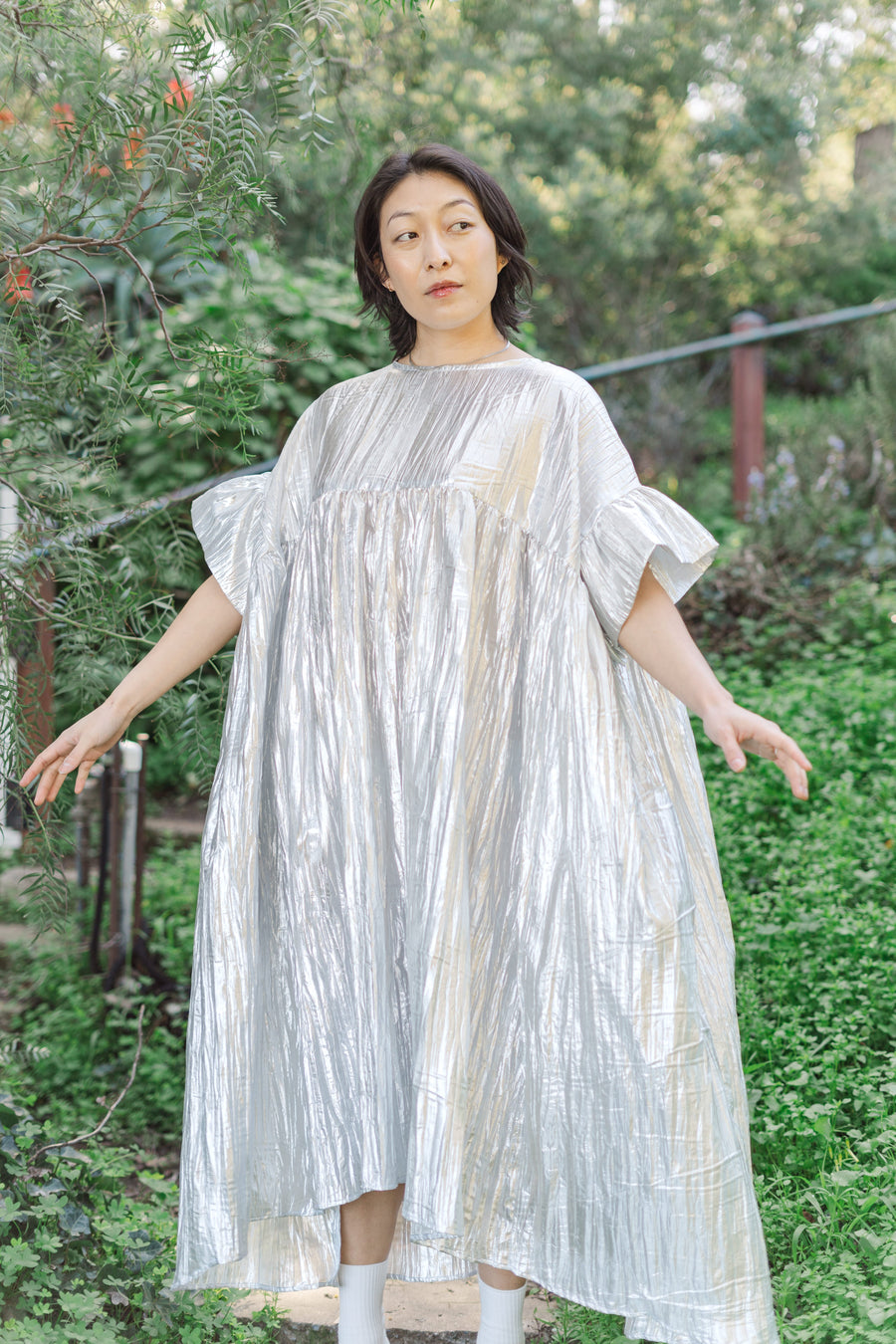 Full-length outdoor portrait of a model in a lush green garden setting wearing the silver metallic crinkle lamé mushroom dress showing its slight high-low hem.