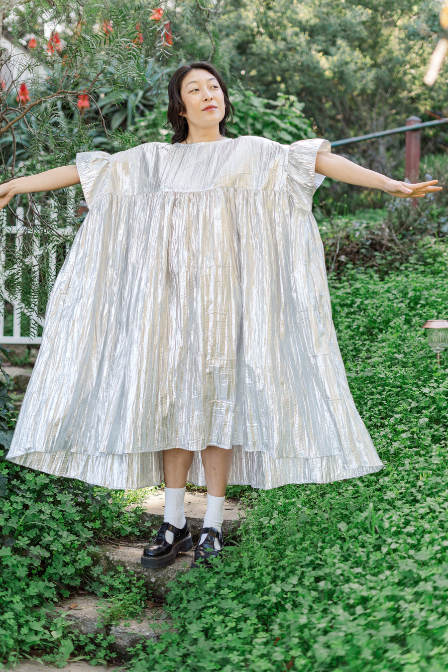 Full-length photo of a model standing in a garden with arms extended to demonstrate the dramatic floaty volume and gathered silhouette of the Silver Metallic Mushroom Dress.
