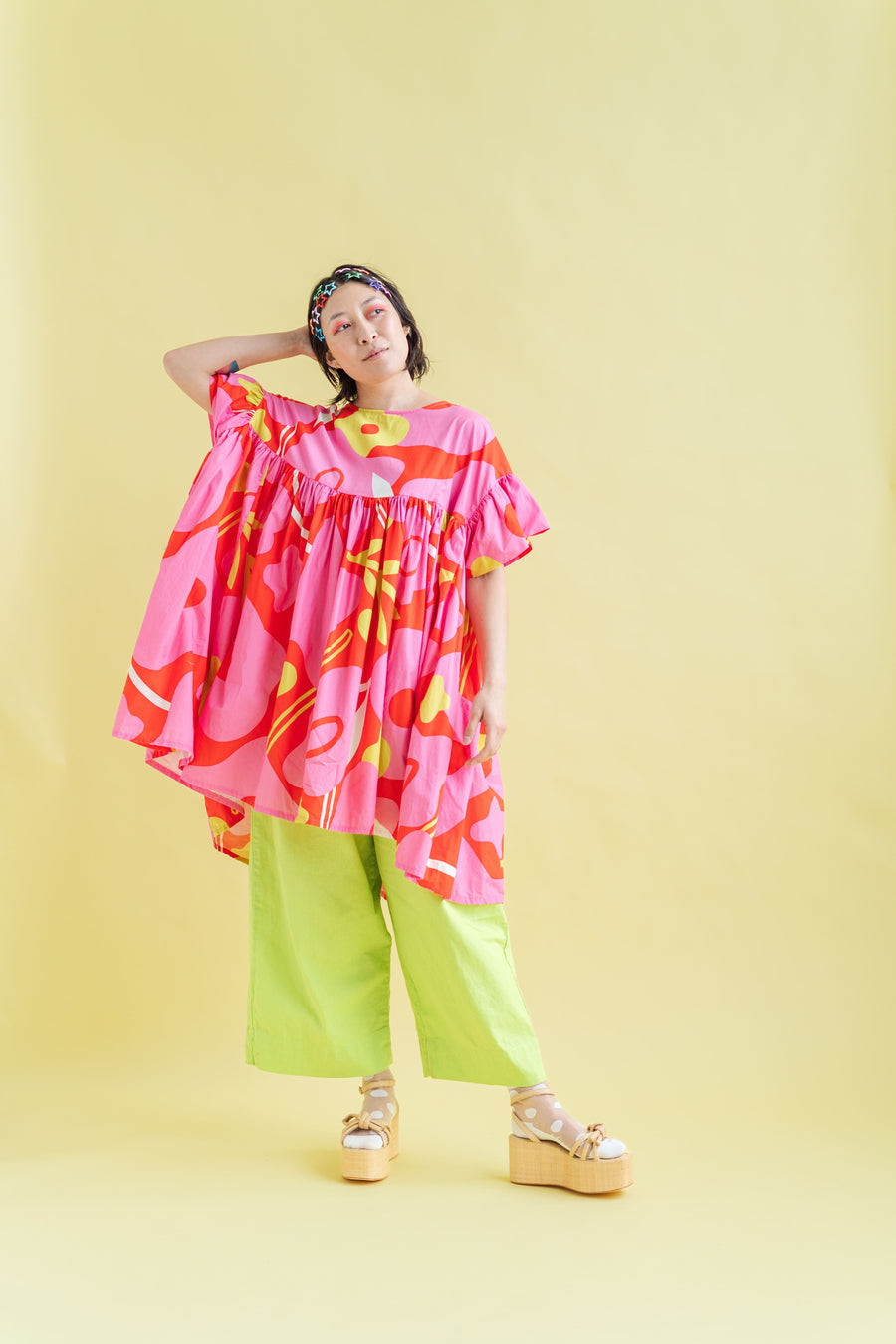 Full-body shot of a model wearing limeade green wide-leg linen pants layered under an oversized patterned tunic against a soft yellow background.