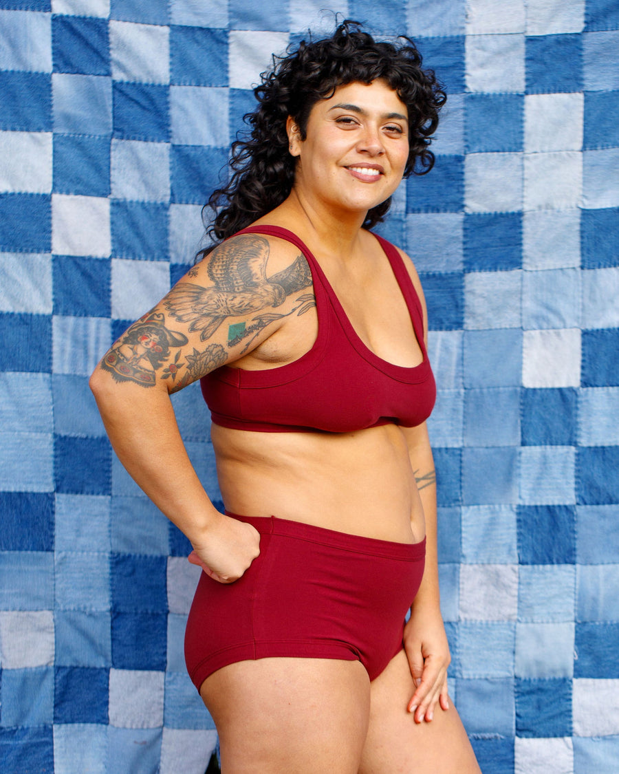 Side profile view of a model with dark curly hair wearing the Tuscan Red organic cotton full brief set against a blue patchwork backdrop.
