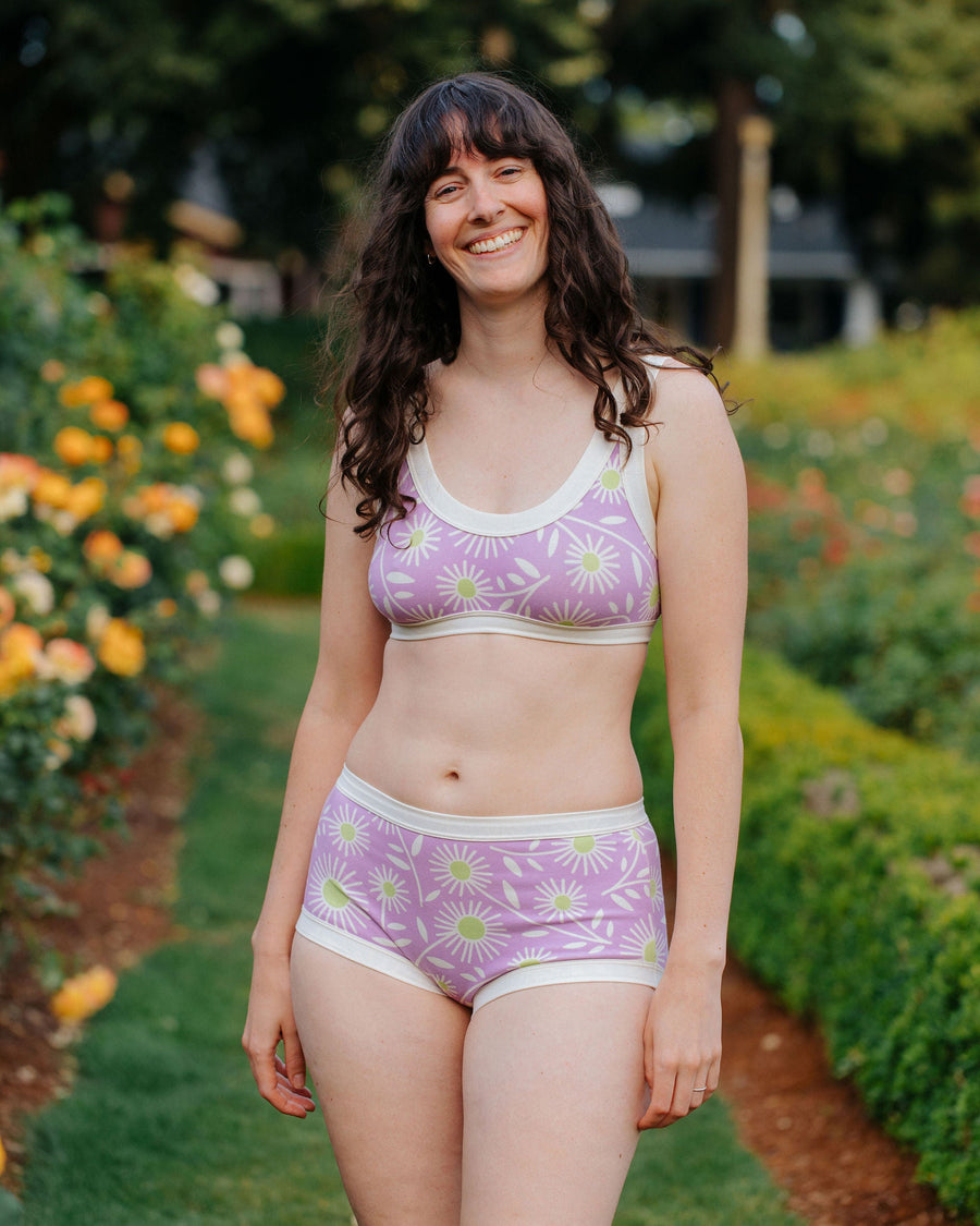 Front view portrait of a smiling model standing in a garden wearing the purple and lime green Delia's Daisy Thunderpants US set.