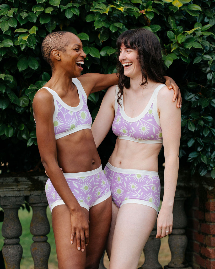 Studio garden portrait of two models embracing while wearing matching purple floral Delia's Daisy organic cotton undies sets.