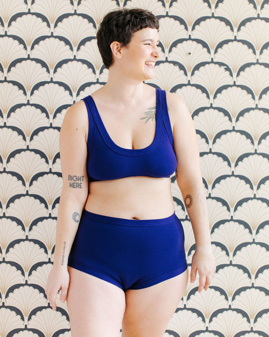 Front view studio portrait of a smiling model with short dark hair wearing the deep blue Indigo Montoya organic cotton brief set against a patterned wallpaper background.