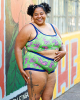 Three-quarter length view of a smiling model with curly hair in pigtails wearing the Electric Tiger matching bra and undies set against a colorful mural wall.