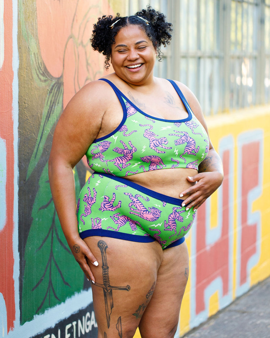 Three-quarter length view of a smiling model with curly hair in pigtails wearing the Electric Tiger matching bra and undies set against a colorful mural wall.