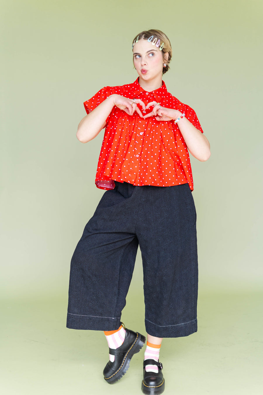 Model making a heart hand gesture wearing the Nettle Studios Black Daisy Denim Mid Easy Pant styled with a red polka dot button-down shirt and chunky black platform shoes.