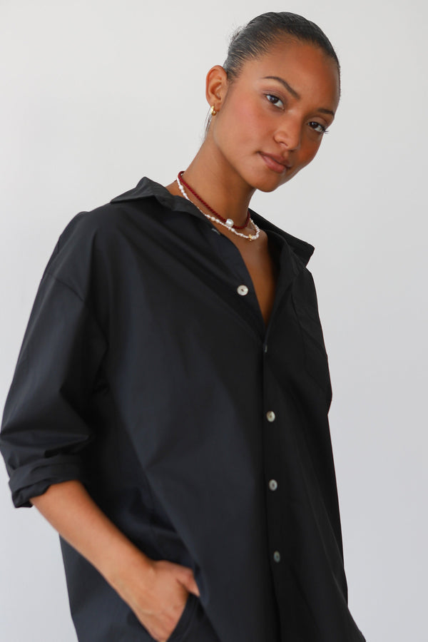 Close-up portrait of a dark-haired model wearing the Be Idle black cotton poplin button-down shirt styled with layered pearl necklaces.