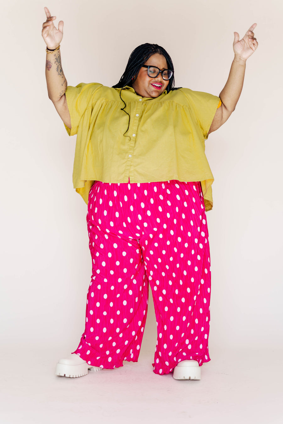 Full-length portrait of a plus-size model raising her hands up while wearing hot pink polka dot plissé flare pants and a yellow button-up top.