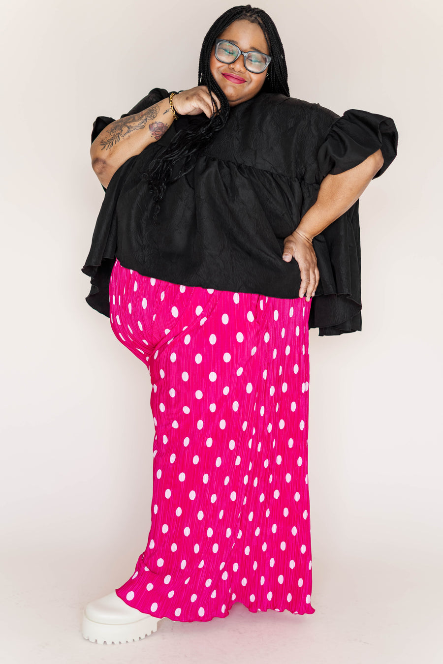 Side profile portrait of a plus-size model wearing hot pink polka dot pleated wide-leg pants styled with an oversized black tiered top.