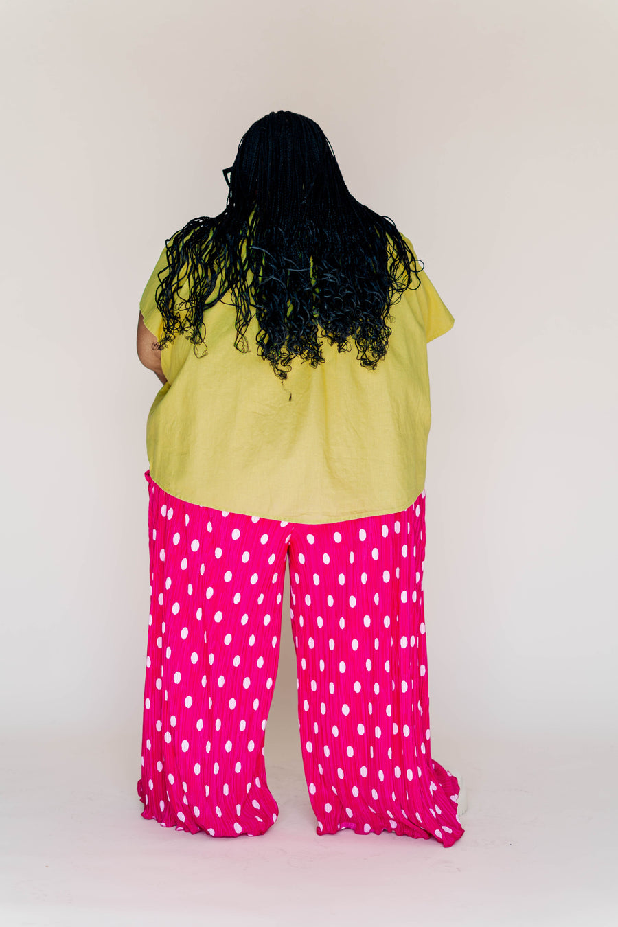 Rear view of a plus-size model wearing hot pink polka dot pleated wide-leg pants paired with a yellow swing top.