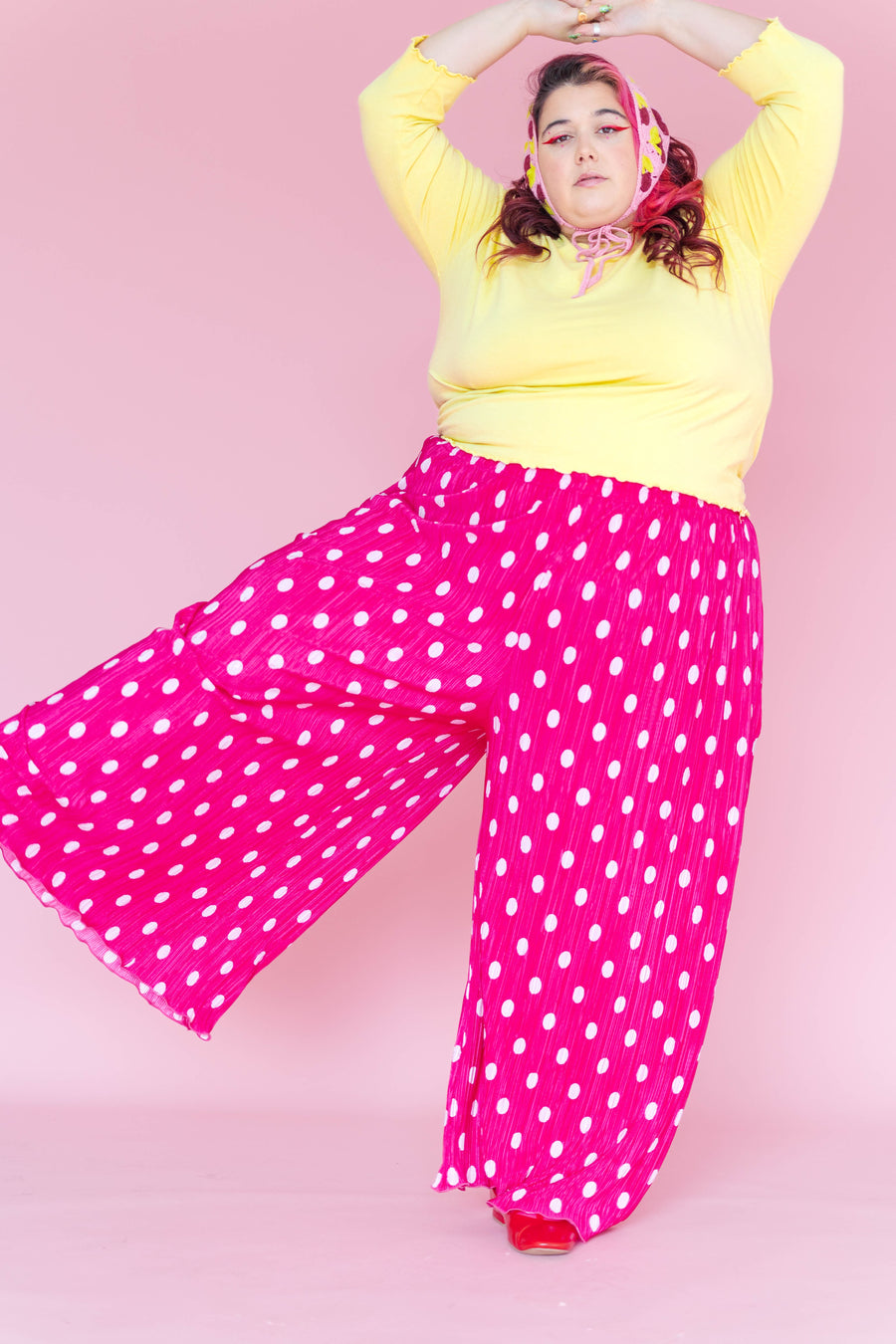 Model kicking her leg out against a pink background while wearing bright pink polka dot pleated flare pants paired with a yellow top.