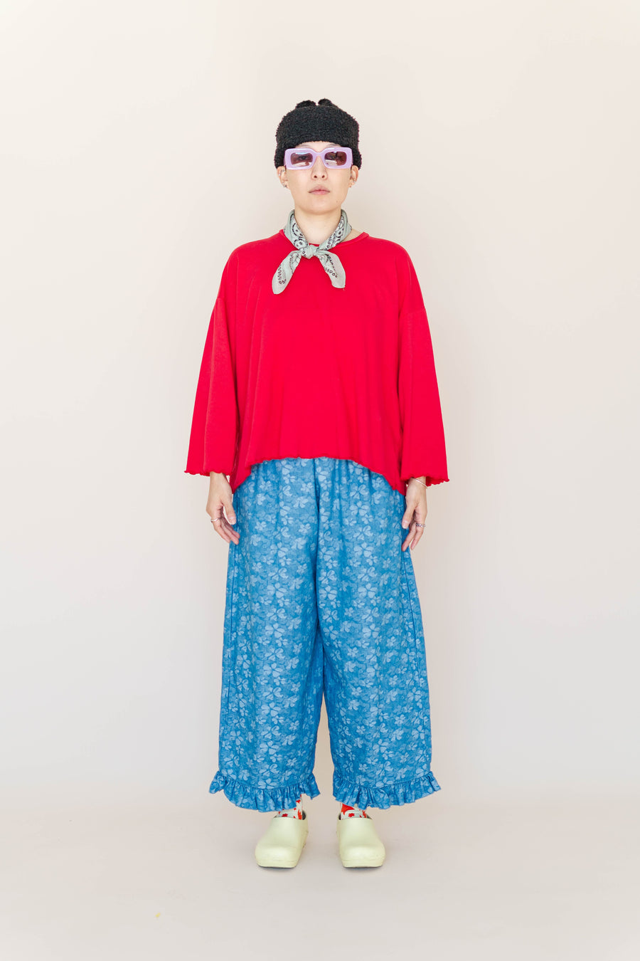 Full-length portrait of model Madison wearing the Light Floral Chambray Ruffle Pant styled with a red long-sleeve top, beanie, bandana, and yellow clogs.