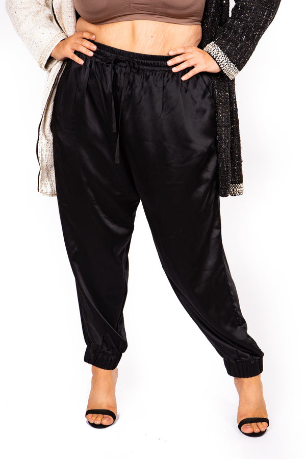A model wearing black satin jogger pants with an elastic drawstring waist and cuffed ankles, styled with a brown crop top, a black cardigan, and black heeled sandals against a white background.