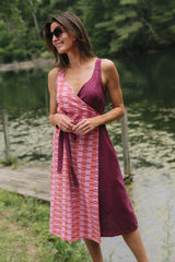 Full-length front view of a model wearing the knee-length Rove wrap dress in Hibiscus styled with sunglasses and hoop earrings by a wooden dock.