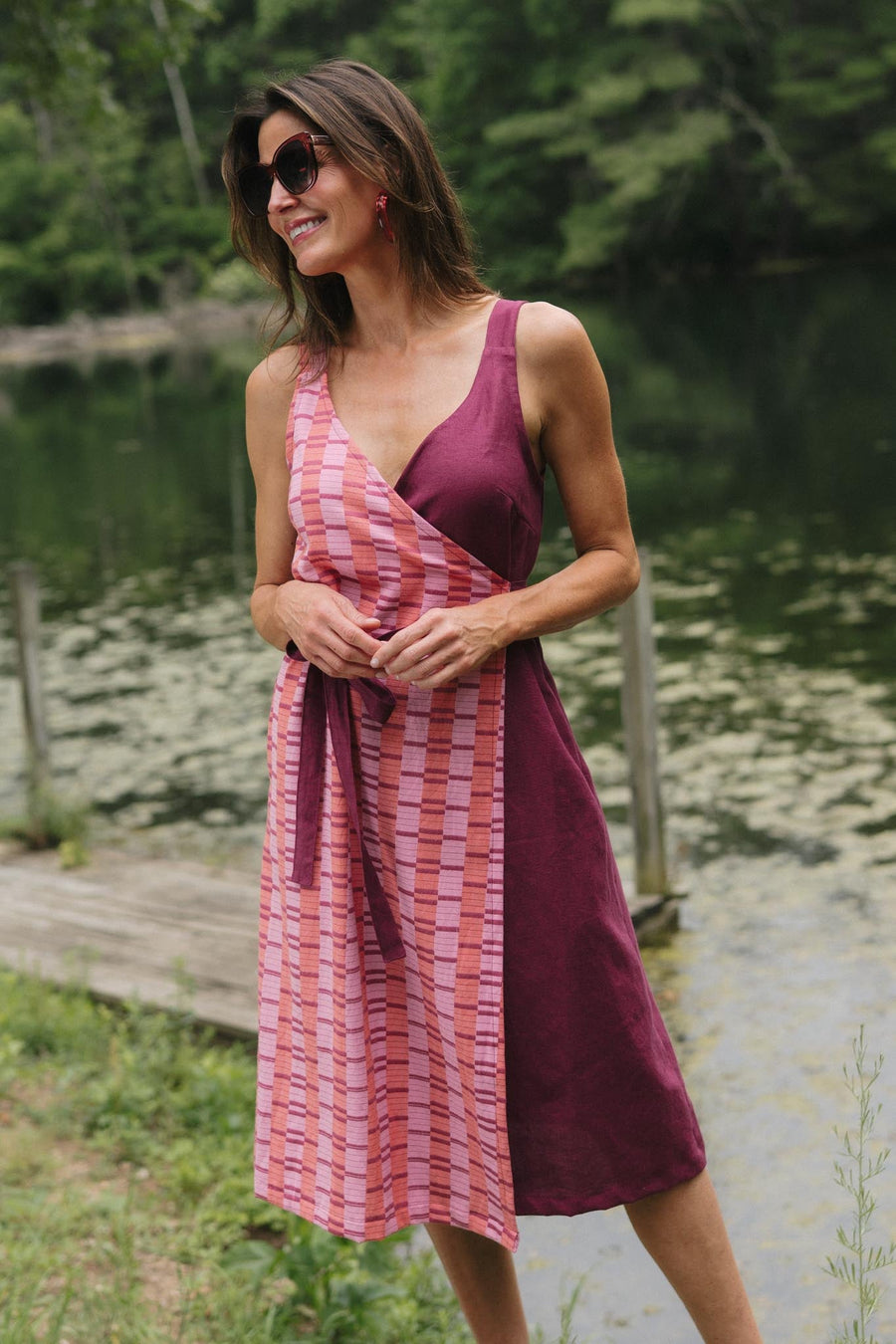 Full-length front view of a model wearing the knee-length Rove wrap dress in Hibiscus styled with sunglasses and hoop earrings by a wooden dock.