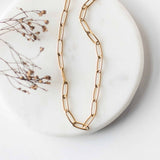Flat lay view of the gold-plated paperclip chain Elle Necklace by Sela Designs displayed on a white marble tray with dried florals.