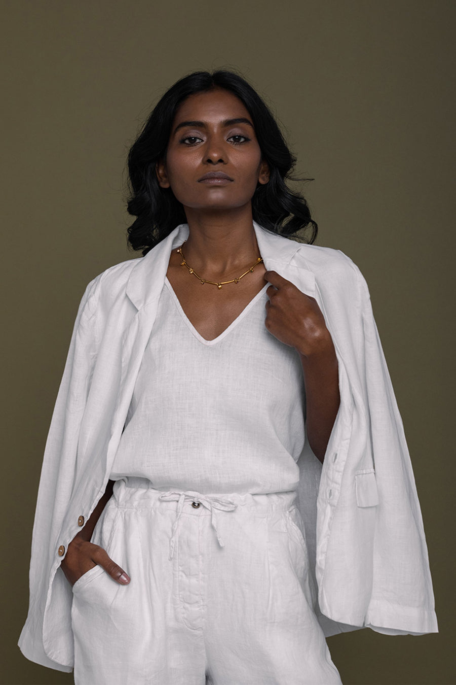 A model wearing the white Reistor hemp blazer draped casually over her shoulders, paired with a matching white V-neck top and draw-string linen trousers against an olive green background.