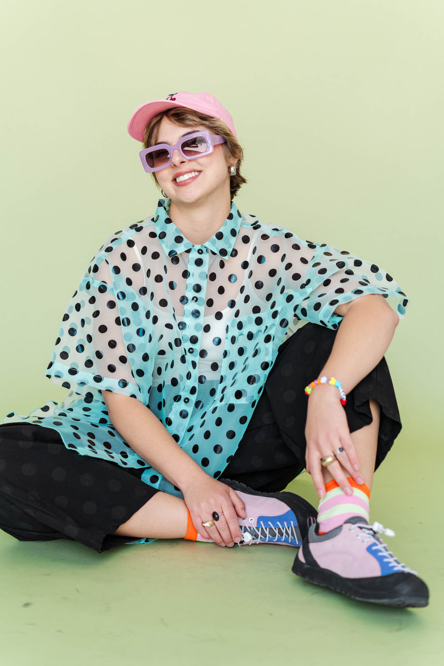 Model seated cross-legged wearing the sheer mint and black polka dot Tulip Top by Nettle Studios. The image emphasizes the short, relaxed sleeves and oversized, airy volume of the size-inclusive organza blouse.
