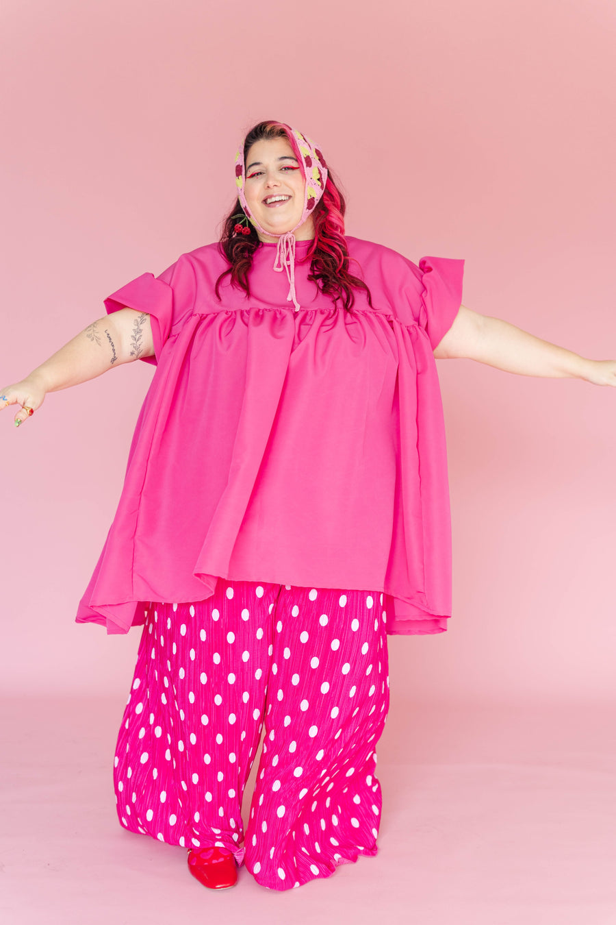 Full-body shot of a model with open arms wearing hot pink polka dot pleated flare pants under a matching hot pink voluminous top.