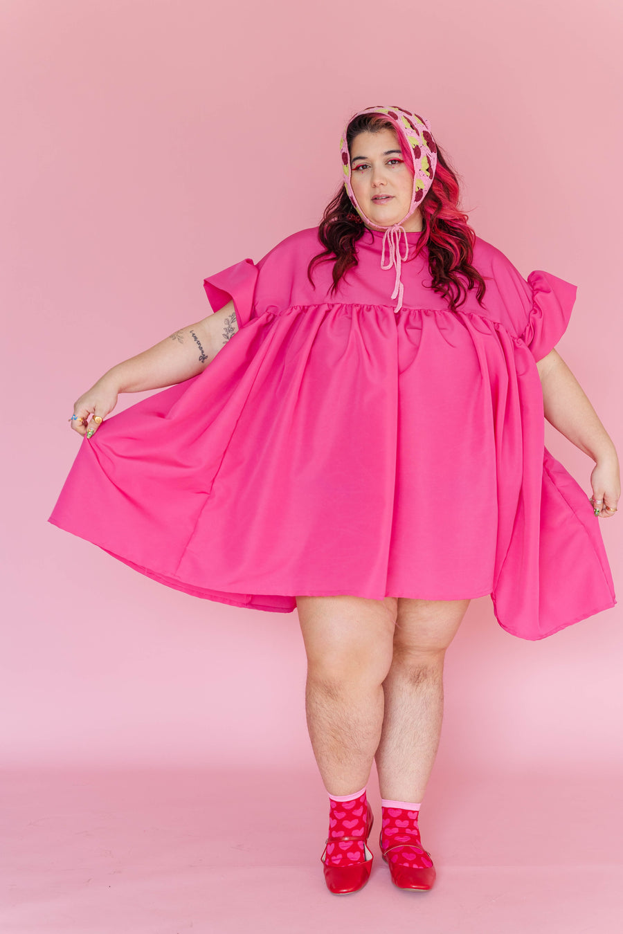Full-length portrait of a model holding the flared hem of the hot pink oversized Mini Mushroom Dress.