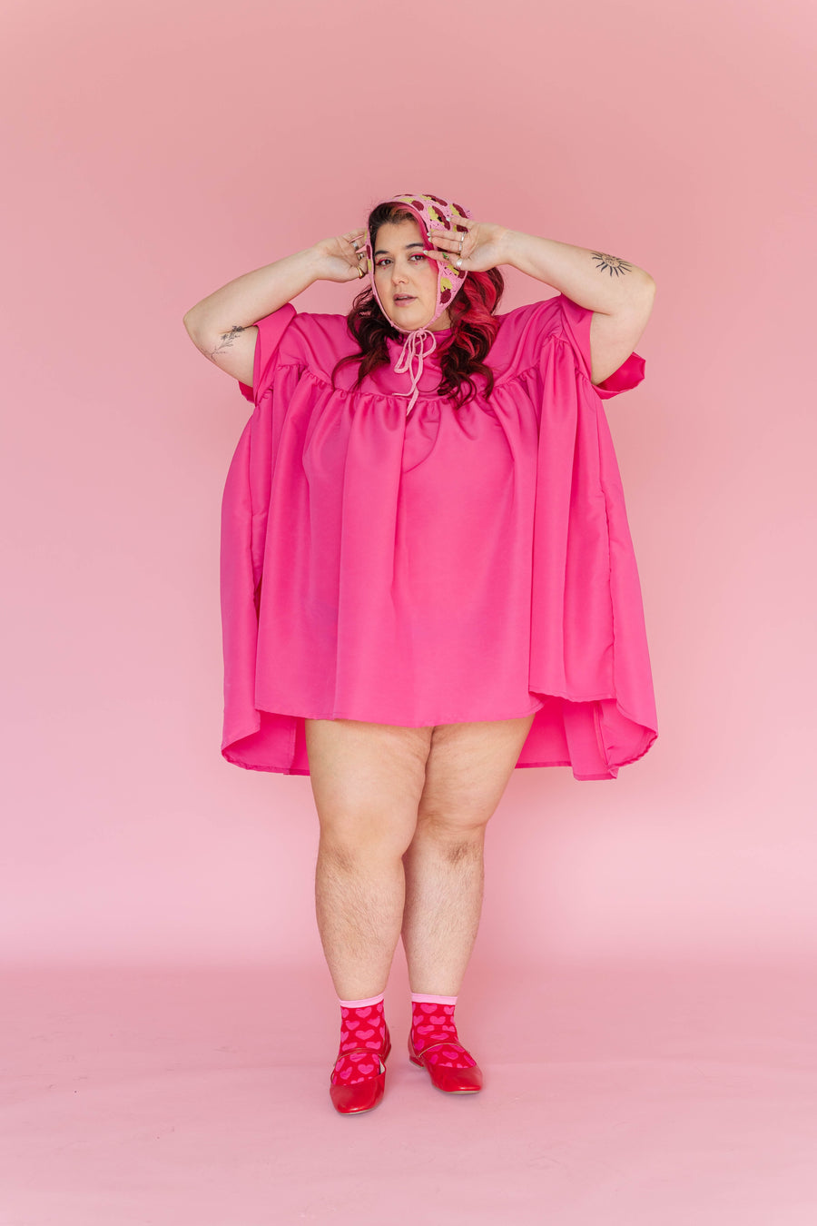 Front view of a model with hands on her head bonnet wearing the hot pink high-low Mini Mushroom Dress by Nettle Studios.