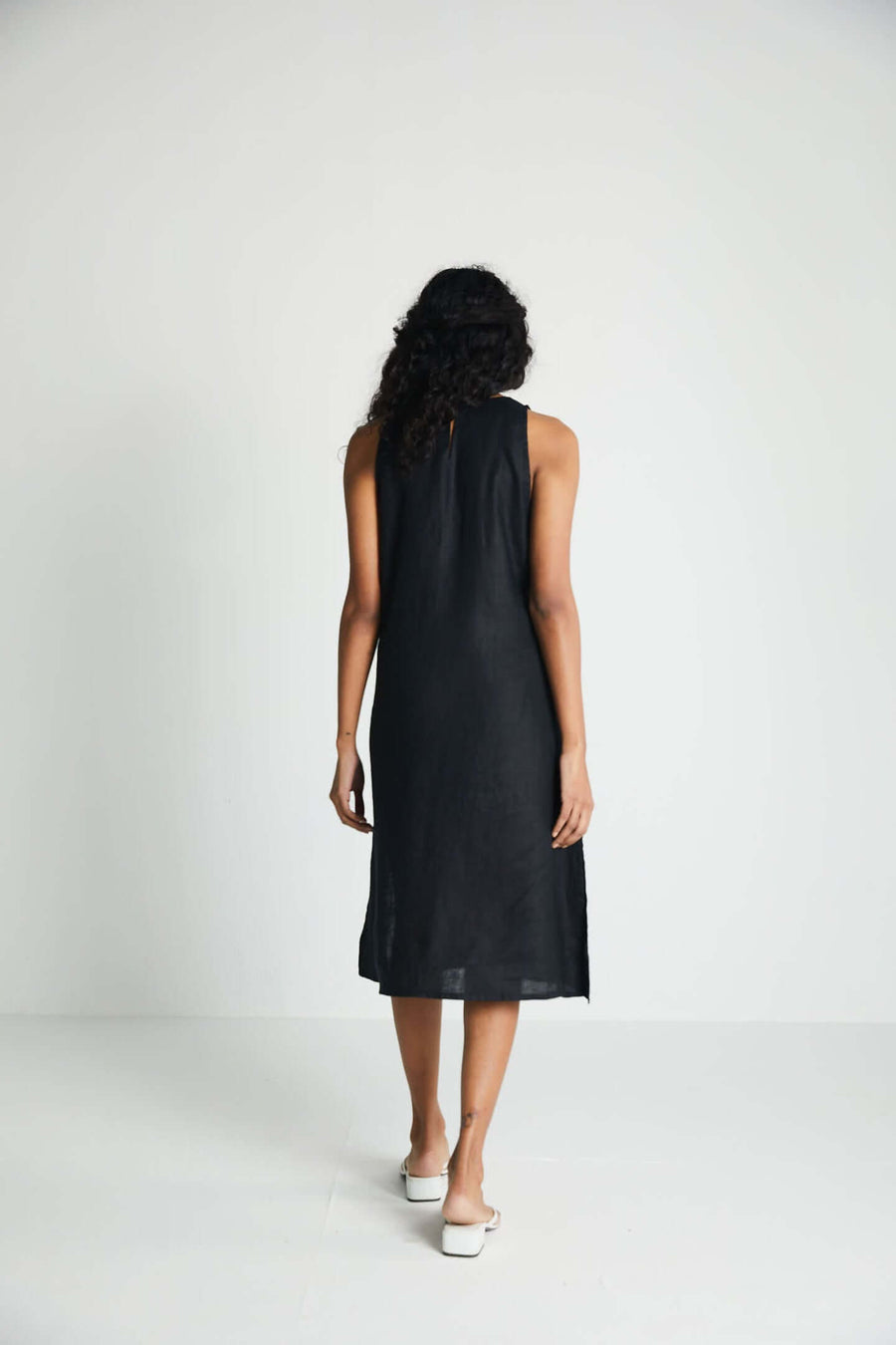 Back view of a model walking in the black sleeveless midi dress, demonstrating the clean fallback drape and high-quality sustainable hemp weave.