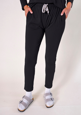Front cropped view of a model wearing black Poplinen Toni Pajama Pants with hands in side pockets, featuring a grey drawstring and elastic waistband.