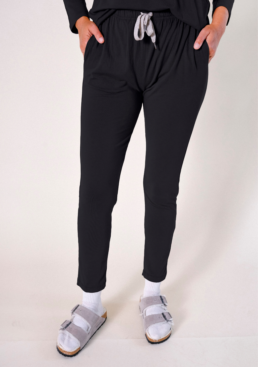 Front cropped view of a model wearing black Poplinen Toni Pajama Pants with hands in side pockets, featuring a grey drawstring and elastic waistband.