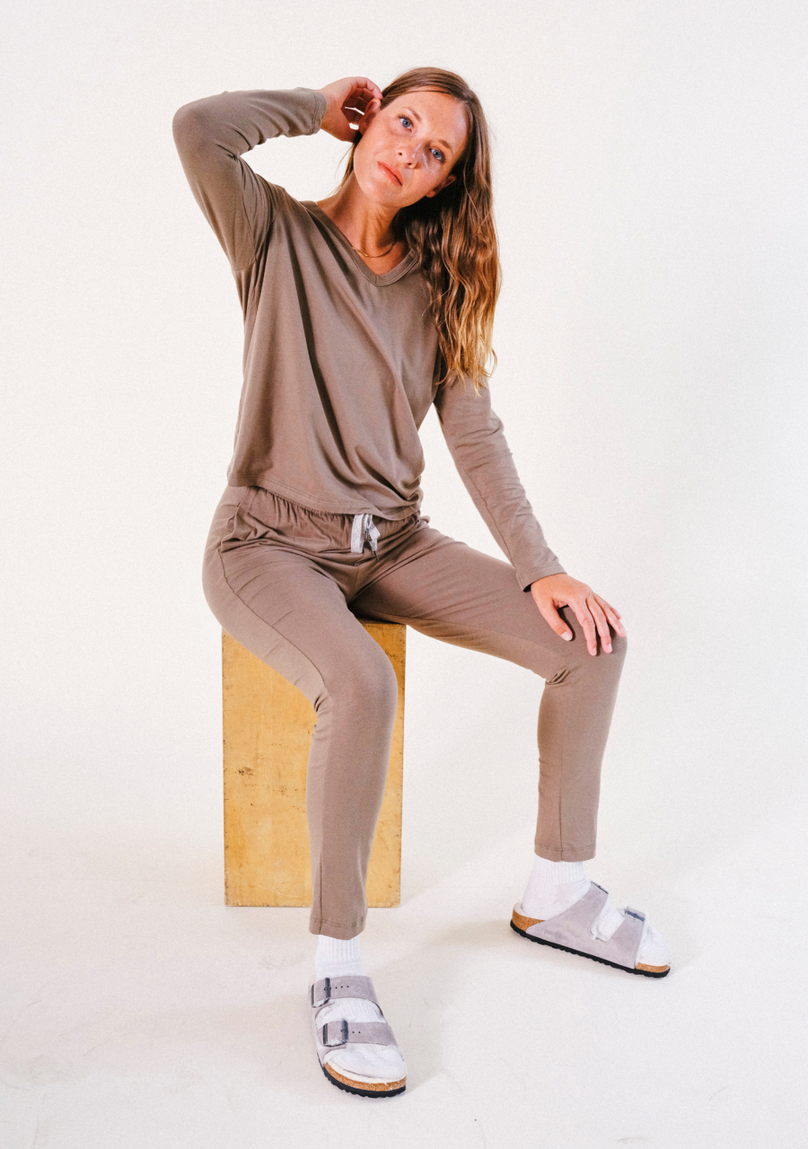 Full-body view of a model sitting on a wooden block wearing the olive-brown Poplinen Toni Pajama set.