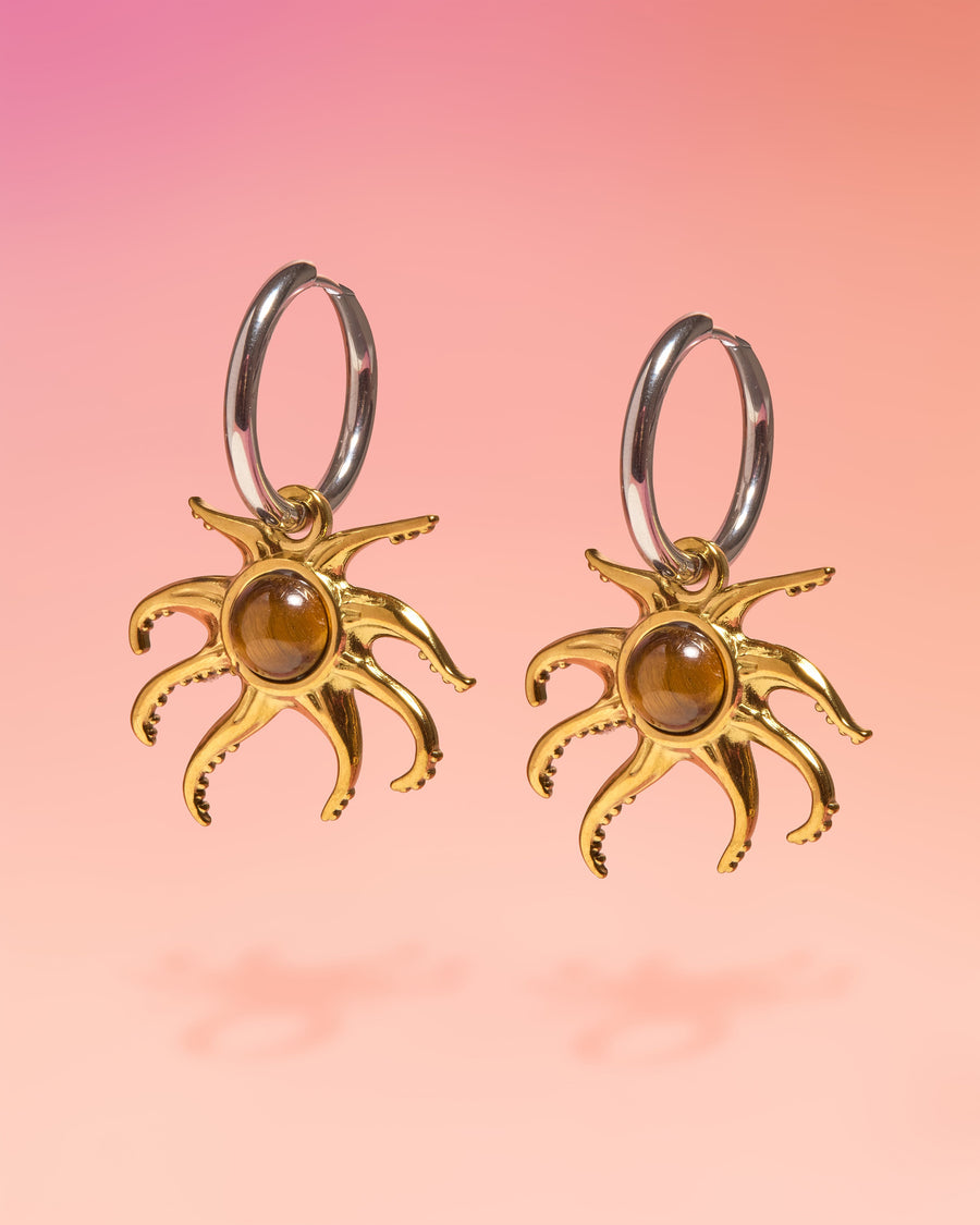 Tribe and Temple Octopus Tiger Eye Hoop Earrings featuring silver surgical stainless steel hoops with a dangling gold-plated octopus charm. The center of each octopus features a round, polished brown tiger's eye stone, set against a soft pink and orange gradient studio background.