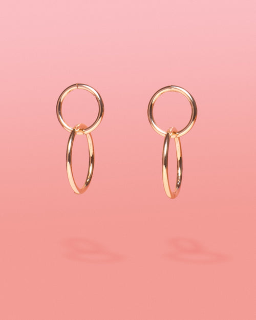 Tribe and Temple Chai Gold 2 Tier Hoops featuring a pair of 18k gold-plated brass double-hoop earrings with interlocking vertical circles on a solid soft pink gradient background.