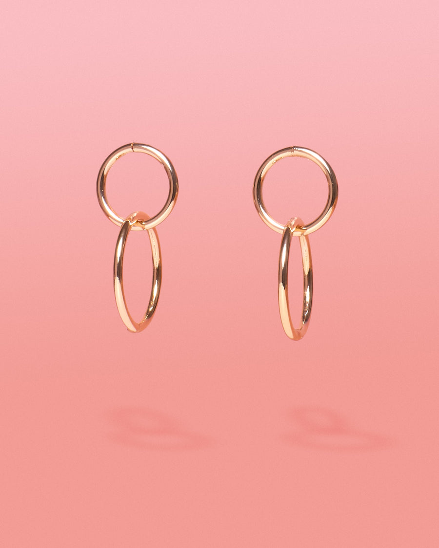 Tribe and Temple Chai Gold 2 Tier Hoops featuring a pair of 18k gold-plated brass double-hoop earrings with interlocking vertical circles on a solid soft pink gradient background.